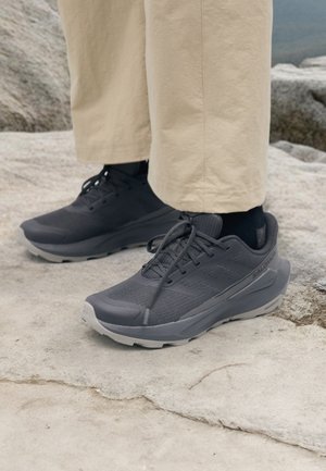 Persoon die zwarte sportschoenen met veters en beige broek draagt, staat op een rotsachtige ondergrond buiten.
