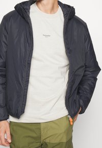 Marineblaues Puffermantel mit Reißverschluss und Kapuze, getragen über einem hellgrauen T-Shirt mit kleinem Aufdruck, kombiniert mit olivgrünen Hosen.
