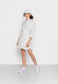 Striped shirt dress with a tie belt, in light colors. Paired with lavender high-top sneakers and a pastel bucket hat. Smooth fabric texture.