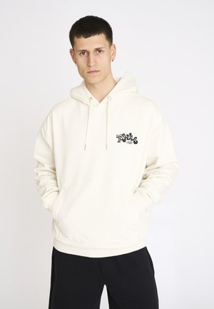 Mand iført en hvid hættetrøje med teksten "Socials Souls" på brystet, stående med hænderne i lommerne foran en hvid baggrund.