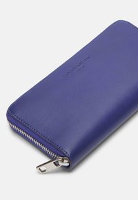 Fioletowy skórzany portfel o gładkiej fakturze, z zamknięciem na zamek i wytłoczonym logo marki. Charakteryzuje się eleganckim, minimalistycznym stylem oraz srebrnymi akcentami.