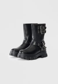Botas de cuero negras hasta el tobillo con suela gruesa, correas de doble hebilla, tachuelas plateadas y un detalle decorativo de cadena en el lateral.