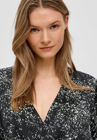 Schwarzes Kleid mit weißen Sprenkeln, V-Ausschnitt und strukturiertem Stoff, mit lässiger Passform. Die Haare sind wellig und schulterlang.