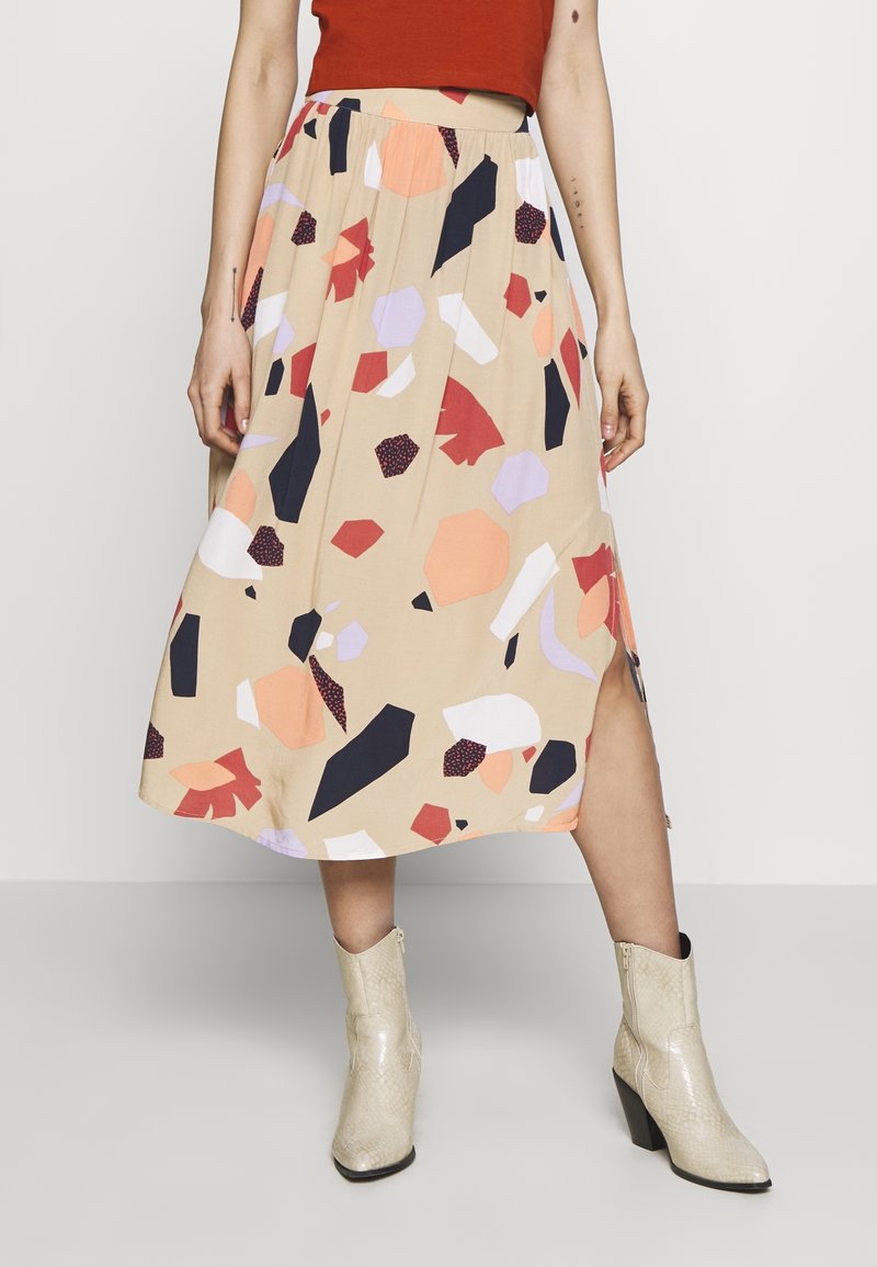 Nainen, jolla on päällä beige midi-hame abstrakteilla monivärisillä muodoilla, ruosteenvärinen crop-top ja vaaleanvalkoiset käärmeennahkakuosiset nilkkasaappaat.