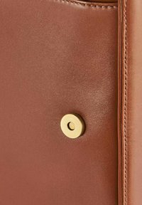Brązowa skórzana torebka z detalami, charakteryzująca się gładką fakturą, starannym przeszyciem oraz okrągłym, złotym zamknięciem magnetycznym.