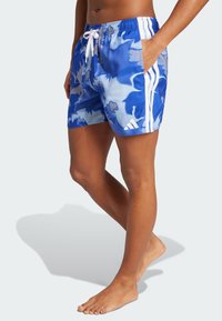 Costumi da bagno blu mimetici con vita elastica e cordoncino. Presentano strisce laterali bianche e tasche. Tessuto liscio e leggero.