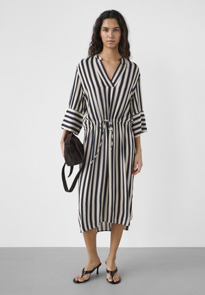 Žena v midi šatách s béžovo-čiernymi vertikálnymi pruhmi a 3/4 rukávmi, drží čiernu tašku a má na nohách čierne sandále na opätku.