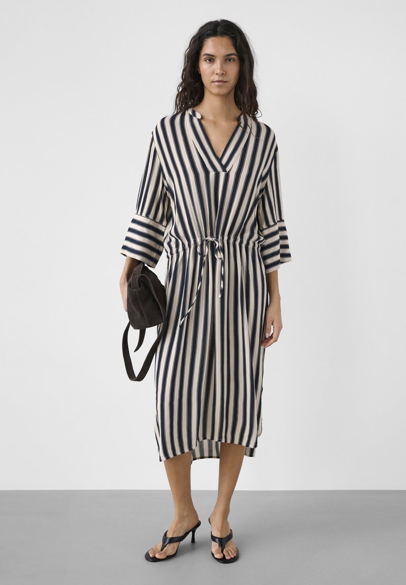 Femme portant une robe midi à rayures verticales beige et noir avec des manches trois-quarts, tenant un sac noir, et portant des sandales à talons noires.