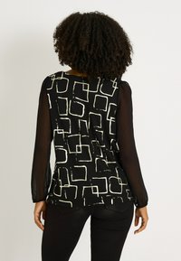 Top negro con mangas largas de tul, con un patrón geométrico de cuadrados blancos. Cuello redondeado, corte relajado y textura suave.