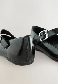 Black patent leather flats with a rounded toe, ankle strap, and silver buckle detailing; smooth, glossy finish and low heel.