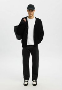 Jaunas vyras su juodu kepurėle, juoda kardiganu, balta marškinėliais, juodais kelnais, juodai-baltomis bateliais, per petį nešantis juodą maišą, stovintis ant baltos fono.