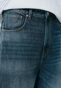 Traperice u tamnom indigo tonu s suptilnom teksturom, s pet džepova i žutim šavovima, koji dodaju kontrast i definiciju.