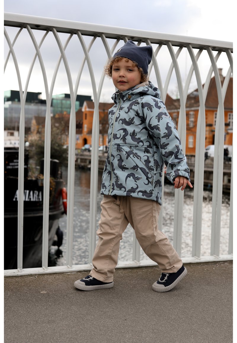 Enfant portant une veste bleu clair avec des motifs animaux foncés, un pantalon beige et des chaussures foncées, debout sur un pont à côté de l'eau.
