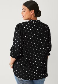 Black polka dot blouse with a ruffled neckline and puffed sleeves, featuring a textured fabric and a relaxed fit. Worn with blue jeans.