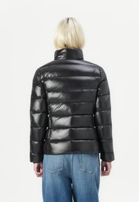 Svart polstret jakke med høy krage, blank finish og horisontal quilting. Bakfra vises elastiske mansjetter og en kortere lengde.