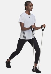 Camiseta atlética blanca con logo negro, combinada con leggings negros texturizados y zapatillas negras. La persona sostiene bandas de resistencia en posición de ejercicio.