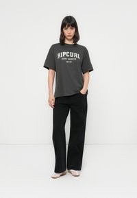 Kvinde iført en mørkegrå Ripcurl Surf Sports T-shirt, sorte brede bukser og lyserødlige flade sko, stående foran en ensfarvet hvid baggrund.