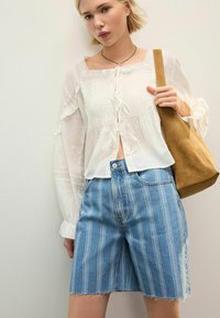 Hvid, let bluse med blonder og snørelukninger, parret med lyseblå stribede denimshorts og en tan ruskinds taske.