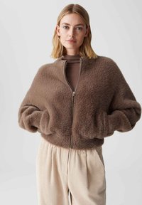 Vrouw met blond haar, gekleed in een bruine fuzzy jas met ritssluiting over een bruine top en lichtbeige corduroy broek, staande tegen een effen achtergrond.