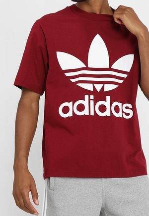 Mężczyzna w czerwonej koszulce Adidas z białym logo i napisem, w połączeniu z jasnoszarymi dresami, stojący na jednolitym tle.