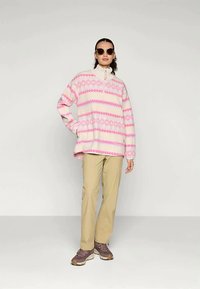Frau mit Sonnenbrille, pink-weiß gemustertem Pullover, beigen Hosen und braunen Sneakers, die mit einer Hand auf der Hüfte vor einem schlichten Hintergrund steht.