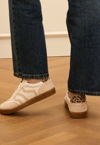 Weiße Sneakers mit beige Wildlederakzenten und Leopardenmuster an der Ferse, ausgestattet mit einer strukturierten braunen Sohle und runden Schnürsenkeln. Kombiniert mit blauen Jeans.