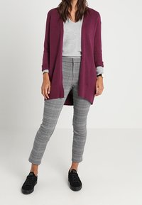 Burgundy kofta över en grå v-ringad topp, kombinerad med grå rutiga byxor och svarta sneakers. Mjuk textur, avslappnad passform.