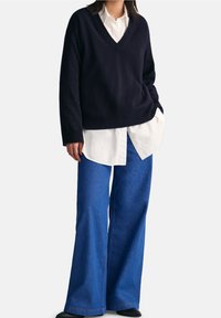 Marineblauer Pullover mit V-Ausschnitt, darüber ein weißes Hemd getragen, kombiniert mit weit geschnittenen blauen Jeans. Der Pullover hat eine lockere Passform und eine weiche Textur.