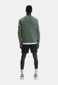 Mężczyzna odwrócony plecami, ubrany w zieloną bluzę, czarne szorty, białe skarpetki i białe buty do biegania, na jednolitym tle.