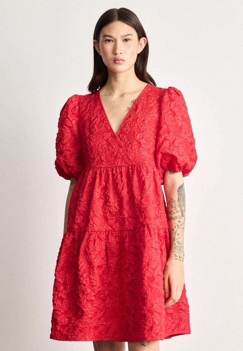 Raudona suknelė su pūstomis rankovėmis, V formos iškirpte, gėlėmis tekstūruotu audiniu, daugiasluoksniu dizainu ir A formos siluetu.