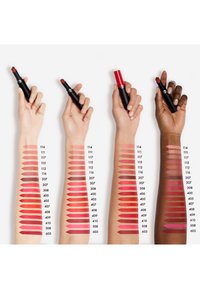 Trzy dłonie trzymają produkty do ust nad próbkami na ramionach, prezentując różne odcienie szminek w kolorach czerwonym, różowym i nude. Szminki mają czarne opakowania.