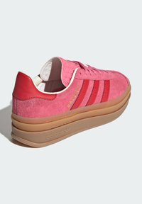 Sapatilhas de camurça cor-de-rosa com acentos vermelhos, atacadores planos e uma sola robusta de borracha bege. Apresenta a marca "Gazelle" em dourado na lateral.