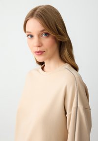Felpa beige con collo rotondo, maniche lunghe e dettaglio metallico lungo la cucitura della spalla, realizzata in tessuto liscio e morbido.