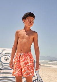 Shorts de bain orange avec motifs de requins blancs et de corail, taille élastique avec cordon de serrage, portés sur une plage de sable avec un parasol rayé.