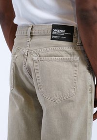 Beige Denim-Jeans mit strukturierter Oberfläche, ausgestattet mit zwei Gesäßtaschen und einem schwarzen Etikett am Bund. Einfaches und funktionales Design.