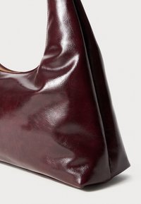 Burgundia nahast käekott, millel on sile, läikiv pind ja iseloomulik ümar kolmnurkne kuju. Elegantselt kujundatud, ilma nähtava riistvaraeta.