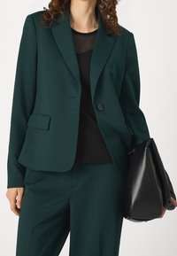 Blazer bleu sarcelle au design structuré, doté d'une fermeture à un bouton et d'une poche. Porté avec un haut noir et tenant un sac noir.