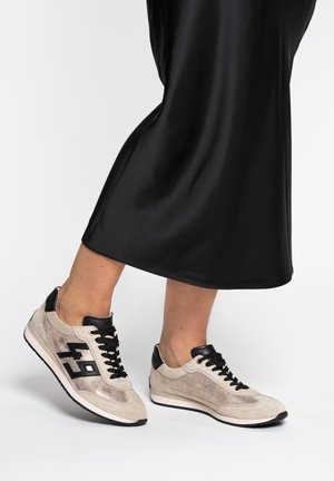 Personne portant des baskets beige et noires à lacets avec des accents noirs et une robe noire, montrant les jambes inférieures sur un fond blanc.