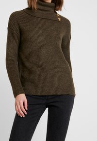 Pull en maille col roulé de couleur vert olive foncé, avec des côtes horizontales, un détail de bouton sur le côté et une coupe décontractée. Fabriqué en matériau texturé.