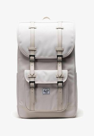 Sac à dos rectangulaire beige clair avec rabat et poche avant fermée par deux sangles à boucle verticales, avec une étiquette de marque Herschel sur la poche.