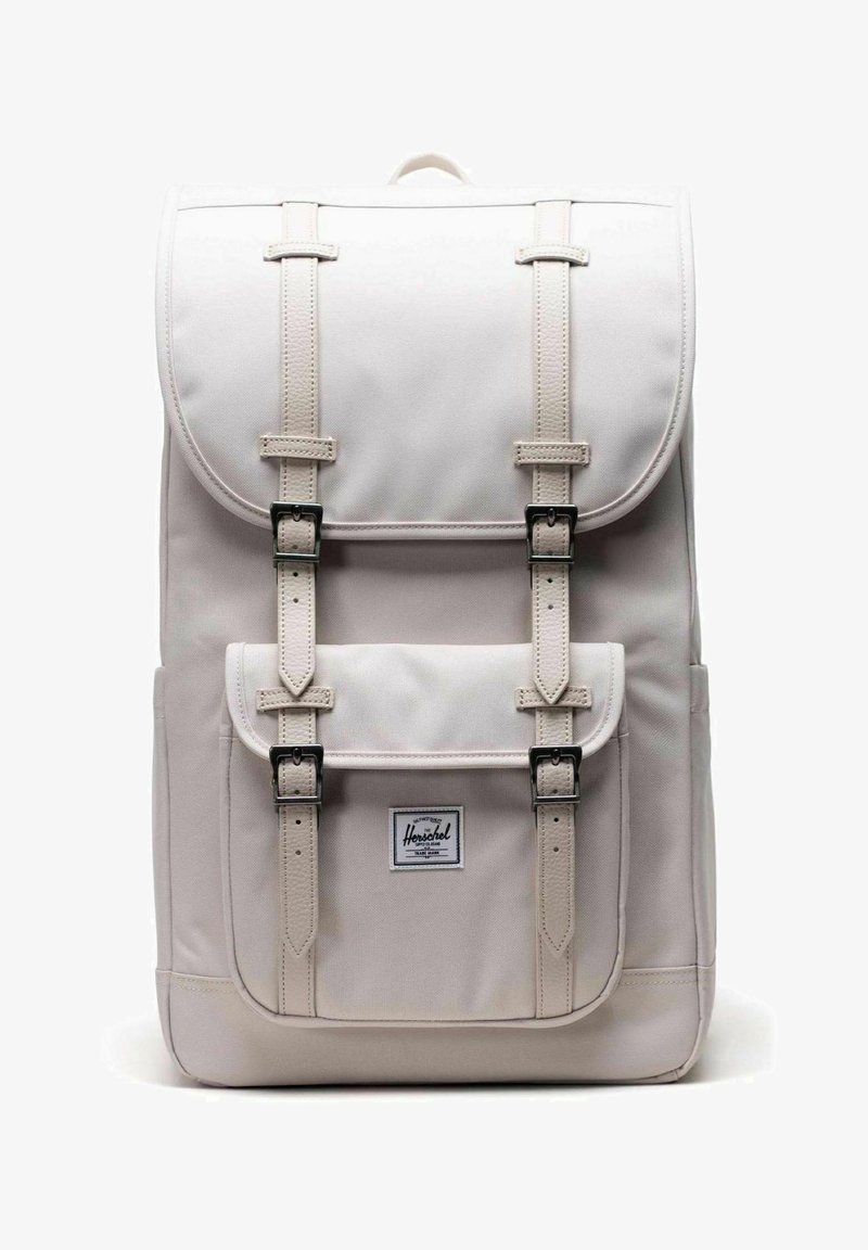 Sac à dos rectangulaire beige clair avec rabat et poche avant fermée par deux sangles à boucle verticales, avec une étiquette de marque Herschel sur la poche.