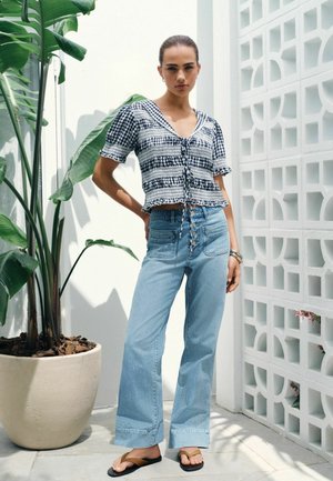 Junge Frau trägt ein blau-weiß kariertes kurzärmeliges Oberteil und hoch taillierte, weit geschnittene Jeans, steht neben einer großen Topfpflanze und einer weißen Wand.