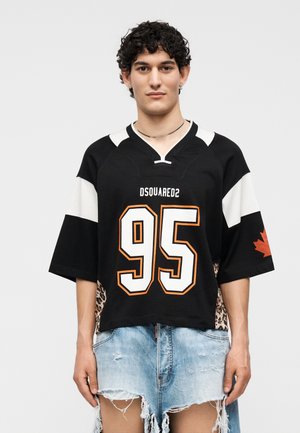 Νέος άνδρας με σγουρά μαλλιά που φοράει μαύρη και λευκή φανέλα DSQUARED2 με αριθμό 95 και ξεβαμμένα ανοιχτά μπλε τζιν σορτς.