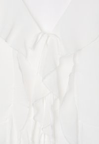 La camicetta bianca presenta uno scollo a V e un design con volant sovrapposti. Realizzata in un tessuto leggero e strutturato, con chiusura a fiocco al collo.