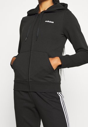 Person mit schwarzem Adidas-Kapuzenpullover mit Fronttaschen und passenden schwarzen Hosen mit weißen Streifen, Hände in den Taschen, Oberkörper sichtbar.