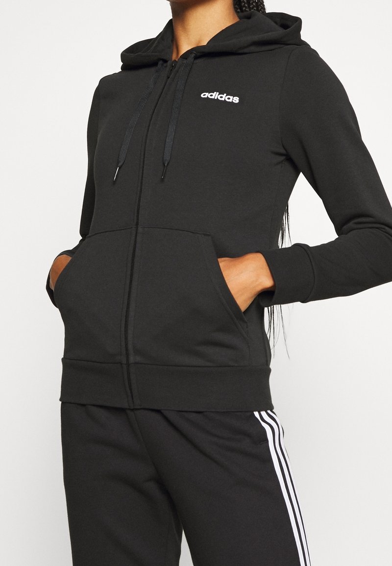 Personne portant un sweat à capuche Adidas noir avec des poches à l'avant et un pantalon noir assorti avec des bandes blanches, les mains dans les poches, haut du corps visible.