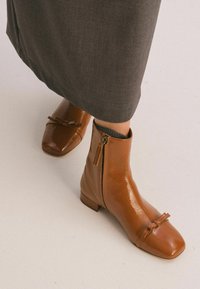 Botines de cuero marrón con puntera cuadrada, un pequeño lazo decorativo y cremallera lateral para el cierre. Llevados con una falda gris.