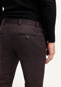 Dunkelburgunderfarbene, gemusterte Hosen mit Slim-Fit, ausgestattet mit zwei Gesäßtaschen und einem schwarzen Knopfdetail. Strukturiertes Gewebe sichtbar.