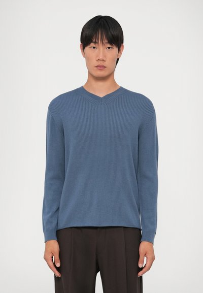 Μπλε ριγέ μακρυμάνικη μπλούζα με σχέδιο v-neck, που διαθέτει υφασμάτινη υφή και ευθεία εσθήτα, φορεμένη με σκούρα παντελόνια.