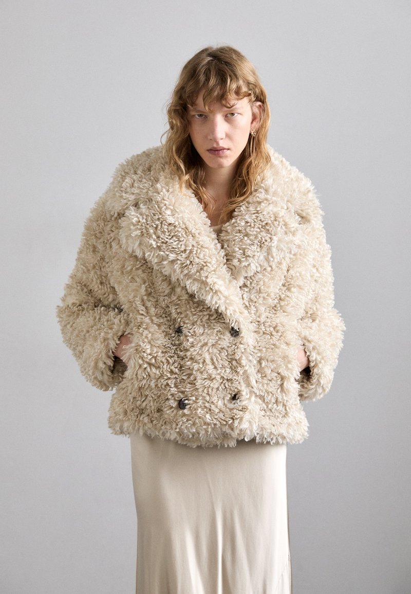 Beige, dubbele-breasted nepbontjas met grote kraag en een getextureerd, fluffy oppervlak. Uitgerust met twee zijzakken en zwarte knopen.